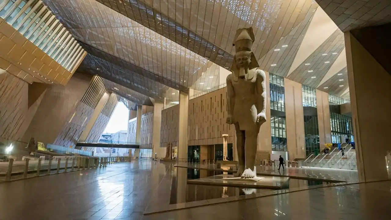 ma foto grandiosa do átrio principal do museu, mostrando a estátua colossal de Ramsés II com a luz do sol entrando pelo teto de vidro, ou uma vista externa do prédio moderno com as Pirâmides ao fundo.