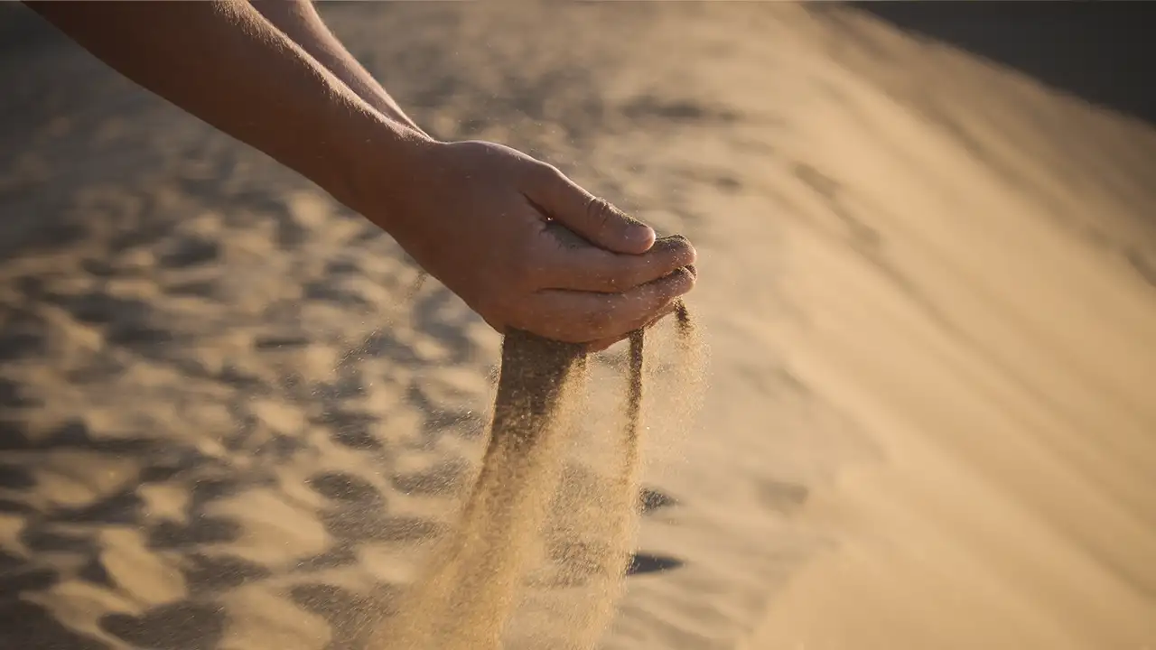 Uma foto do silo da bateria de areia na Finlândia (um grande tanque cilíndrico de aço cinza/marrom) ou uma foto artística de mãos segurando areia quente com painéis solares ao fundo.