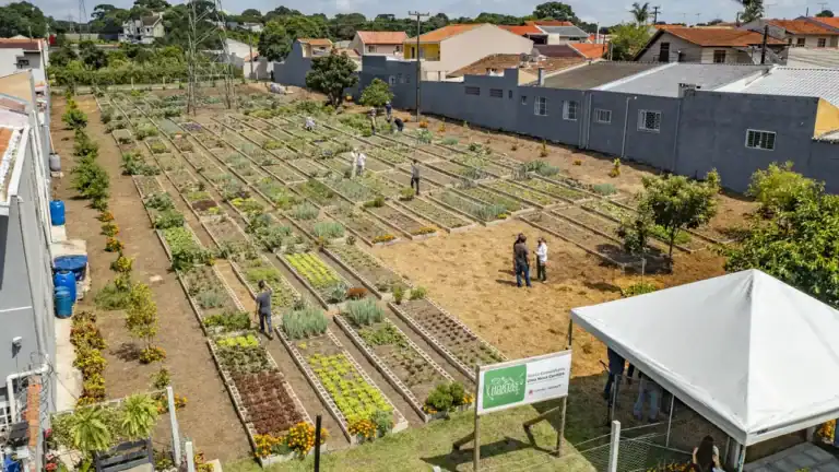 Vista aérea de uma horta urbana comunitária em Curitiba, Brasil, mostrando canteiros verdes de vegetais ao lado de edifícios urbanos, um exemplo de agricultura urbana